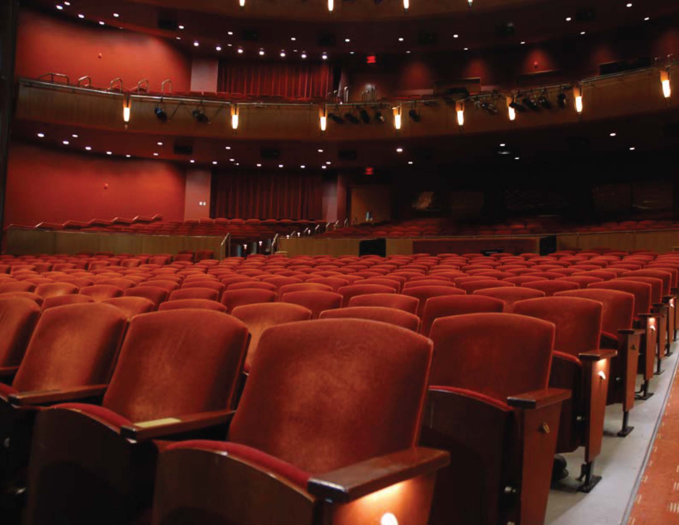 Empty theater auditorium with rows of red seats and balcony lighting.